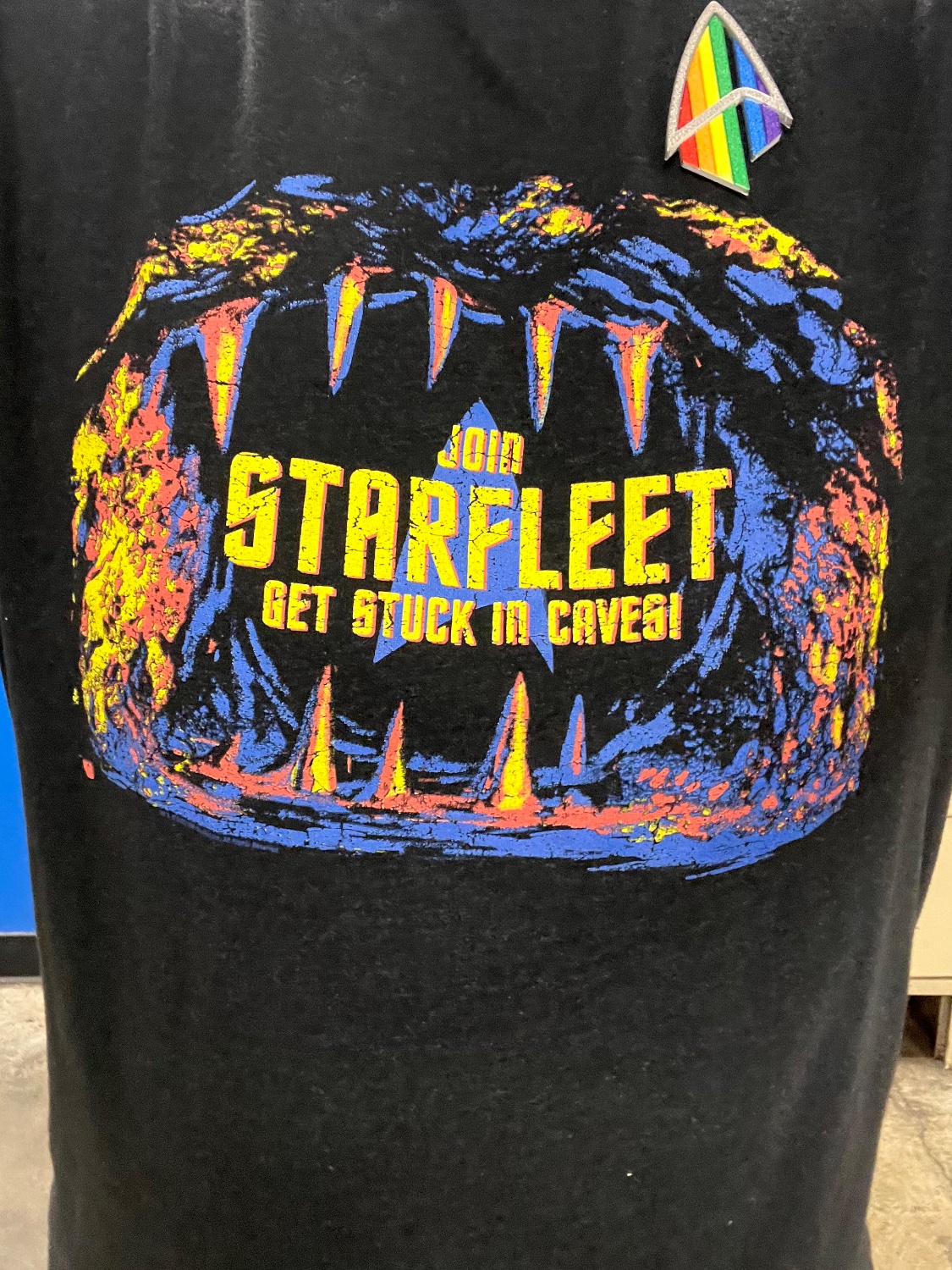 A t-shirt with the cartoon image of a cave that resembles a mouth with sharp fangs. The text says “Join Starfleet Get Stuck In Cave!” The person wearing the shirt is also seen wearing a 3d printed PIC era badge with Pride colors.